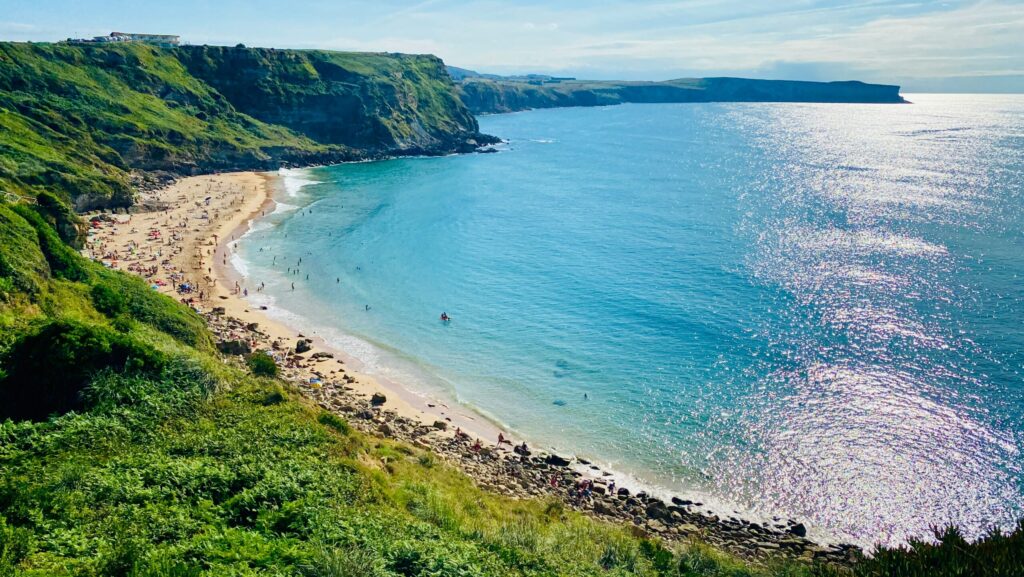Beach at Suances, Spain, with people on the sand and swimming, clear blue flat water, greenery, and cliffs in the background