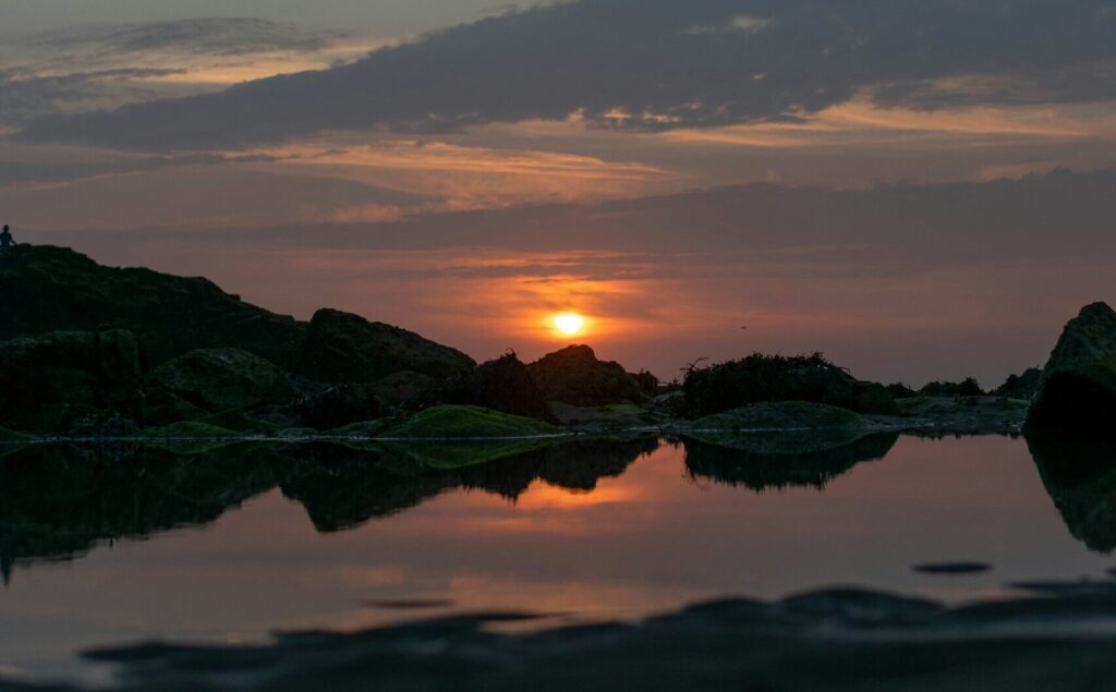 Sunset at Perranporth, UK, with rocks and water in the foreground, cloudy skies, and yellow hues from the sun