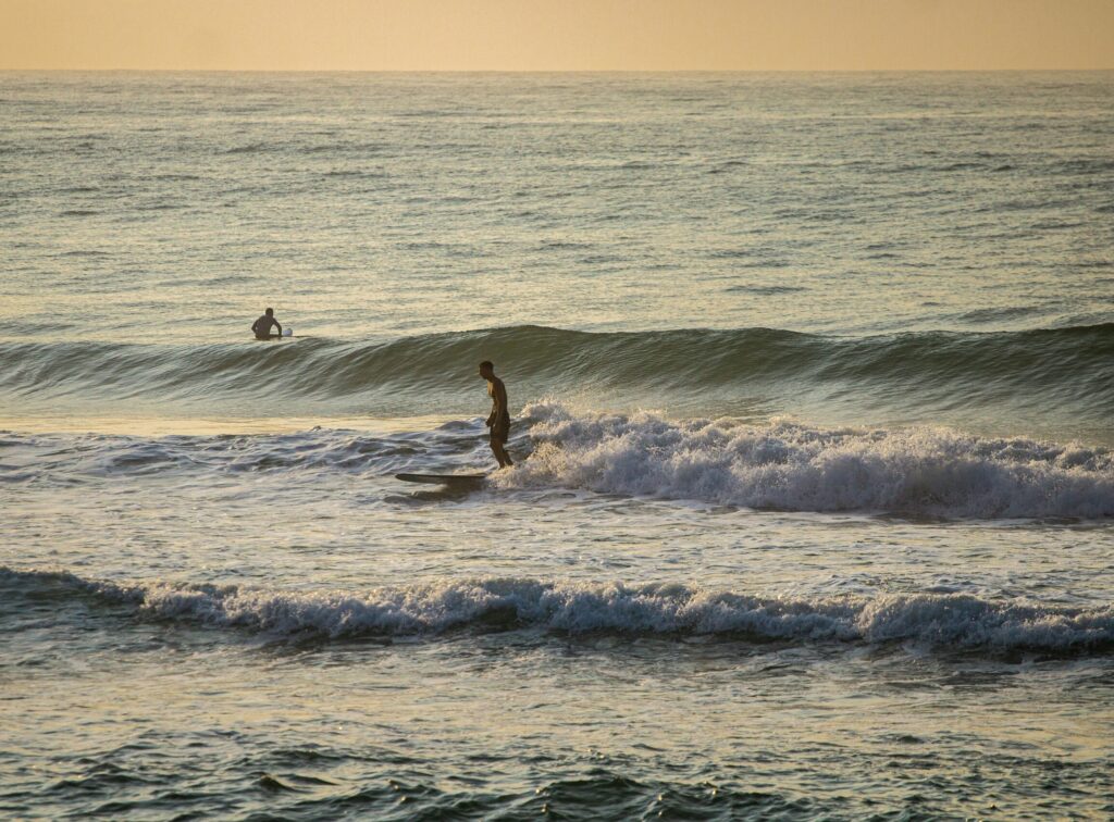 A surfer riding a Malibu on a small wave with a breaking set behind him, Perranporth surf, UK