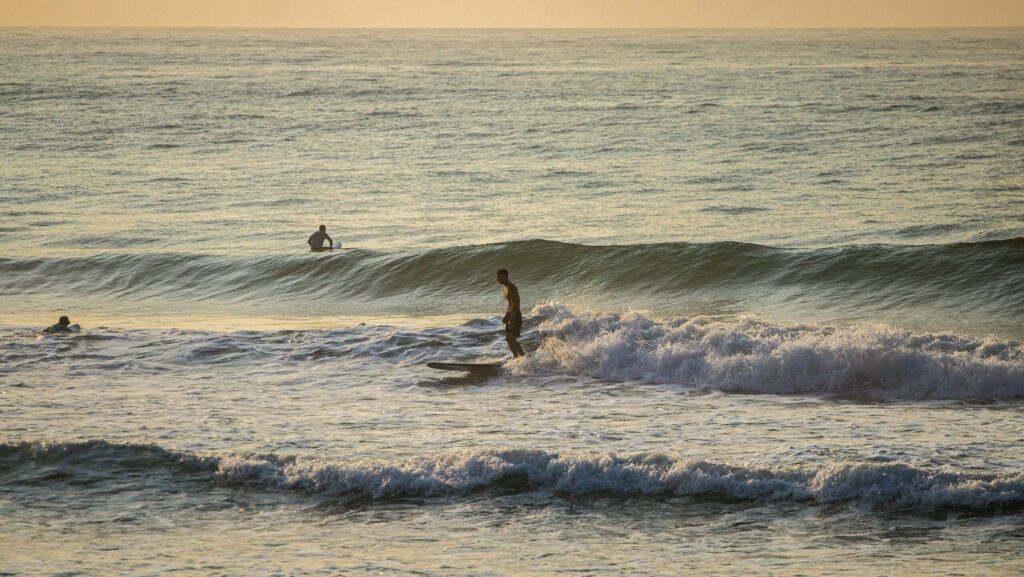 A surfer riding a Malibu on a small wave with a breaking set behind him, Perranporth surf, UK