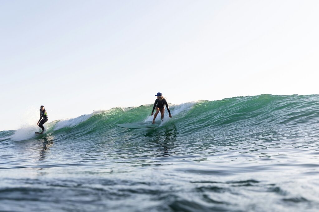 Two surfers on a wave, one dropping in on the other, with clear blue skies and greenish-blue water in Ribadesella Surf, spain