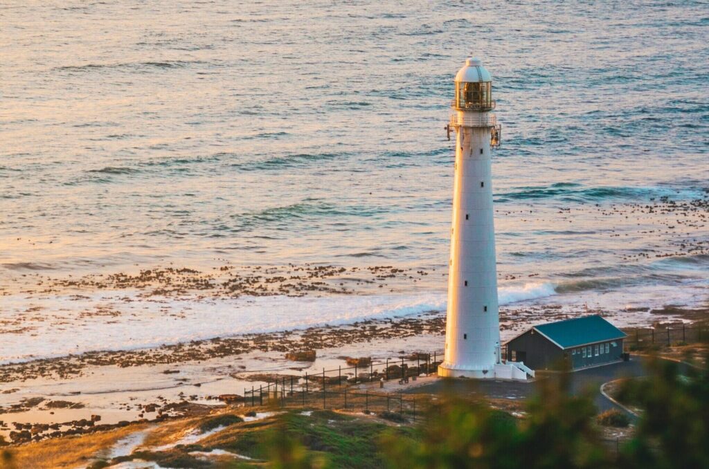 A white lighthouse in Kommetjie, South Africa, with the ocean and sunlight reflecting on the water in the background