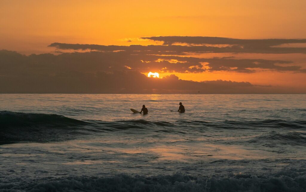 Two surfers sitting in the water at sunset, waiting for the next wave under orange skies in Ribadesella surf, Spain