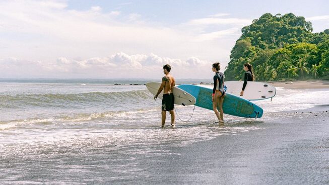Two people with surfboards standing on the shore looking out into the ocean at Damo Surf Camp in Jaco