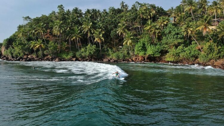 a surfer riding a wave with trees in the background at Surf And Yoga Mirissa Retreat