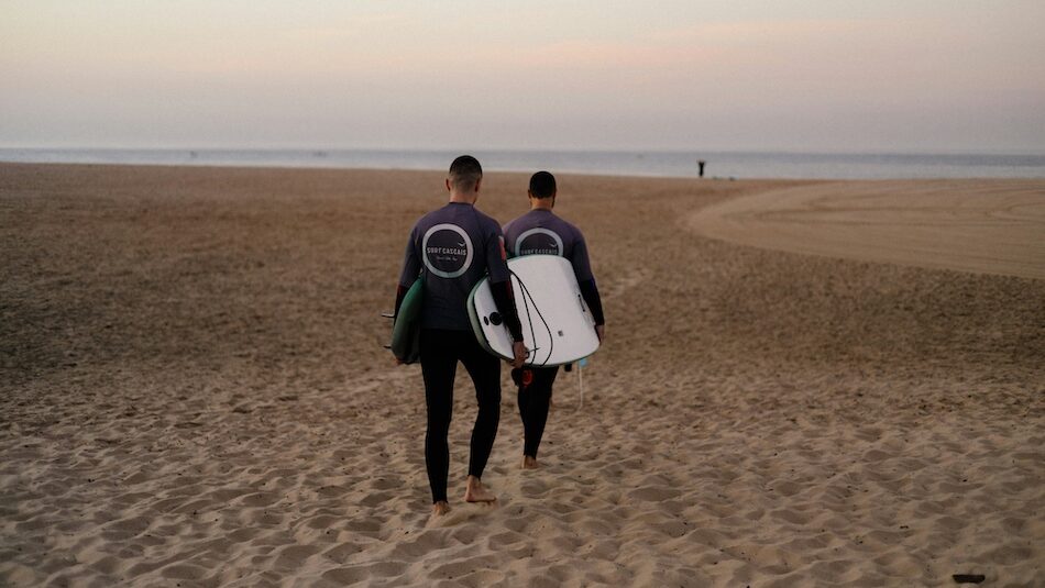 Two people carrying a surfboard on the beach at a surf camp near Lisbon