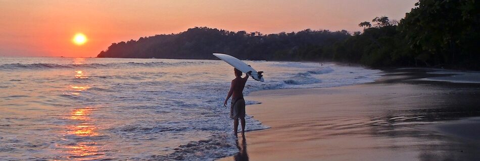 A surfer with the surfboard on his head at sun set at a surf yoga camp Costa Rica