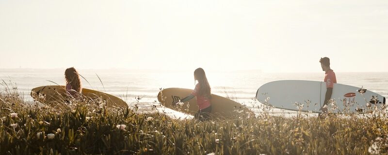 3 people walking with surfboards with the sun behind them thinking Where to surf in Portugal in summer